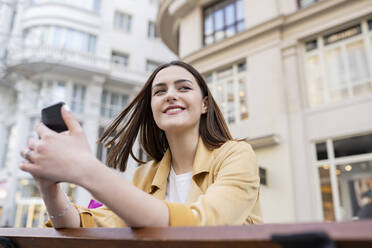 Lächelnde junge Frau, die ihr Smartphone in der Hand hält, während sie vor einem Gebäude wegschaut - JCCMF01405