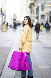 Young woman with shopping bags looking over shoulder while walking on footpath in city - JCCMF01407