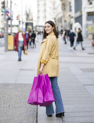 Smiling young woman with magenta colored shopping bags walking on footpath in city - JCCMF01408