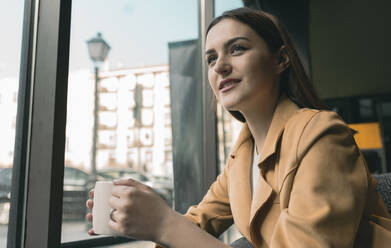 Lächelnde junge Frau mit Kaffeetasse, die durch das Fenster schaut, während sie in einem Café sitzt - JCCMF01415