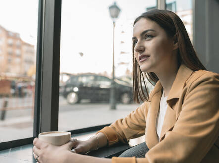 Junge Frau mit Kaffeetasse sitzt am Glasfenster eines Cafés und schaut weg - JCCMF01418