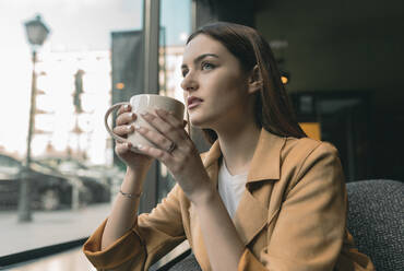 Beautiful woman drinking coffee while looking through window at cafe - JCCMF01419