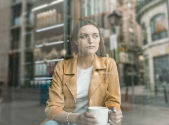 Nachdenkliche junge Frau, die in einem Café sitzt, gesehen durch ein Glasfenster - JCCMF01421