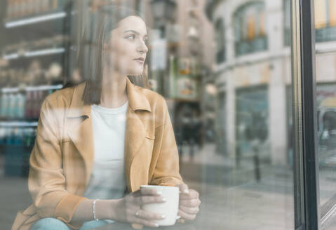 Nachdenkliche junge Frau mit Kaffeetasse, die im Café sitzt und durch das Fenster schaut - JCCMF01423