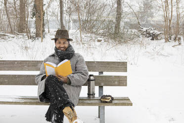 Lächelnder Mann liest ein Buch, während er im Winter auf einer Bank sitzt - AXHF00194