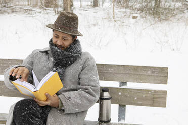 Mid adult man reading book while sitting on bench during winter - AXHF00195