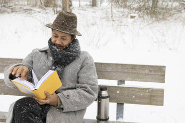 Mittlerer erwachsener Mann, der im Winter auf einer Bank sitzend ein Buch liest - AXHF00195