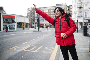 Smiling man holding mobile phone while hailing ride on footpath - ASGF00040