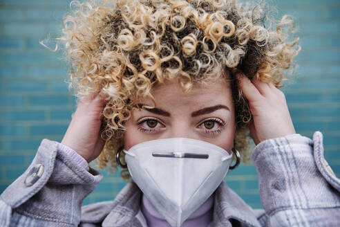 Woman with hand in curly hair wearing protective face mask against wall during COVID-19 - ASGF00049