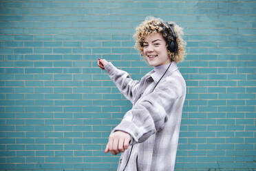 Smiling curly haired woman dancing while listening music through headphones - ASGF00063
