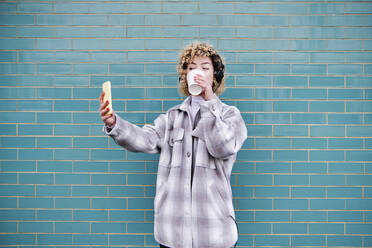 Lockig behaarte Frau, die ein Selfie macht, während sie vor einer Wand Kaffee trinkt - ASGF00069