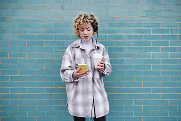 Schöne Frau mit Kaffeetasse, die Musik über Kopfhörer hört, während sie ihr Smartphone vor einer blauen Wand benutzt - ASGF00070