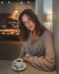 Selbstbewusste junge Frau in eleganter Bluse schaut in die Kamera, während sie sich in einem gemütlichen Café mit einer Tasse Kaffee entspannt - ADSF21266