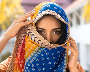 Junge ethnische indische Frau in traditionellem bunten Kopftuch schaut in die Kamera, während sie vor einem unscharfen tropischen Garten steht - ADSF21270