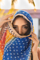 Junge ethnische indische Frau in traditionellem bunten Kopftuch schaut in die Kamera, während sie vor einem unscharfen tropischen Garten steht - ADSF21271
