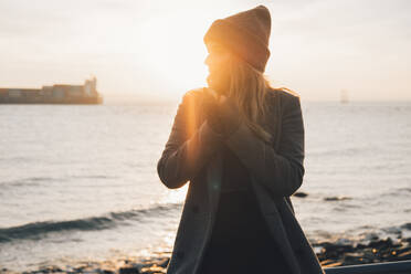 Seitenansicht einer lächelnden jungen Frau in warmem Mantel und Hut, die an der Reling steht und den Sonnenuntergang über dem Meer am Herbstabend bewundert - ADSF21328