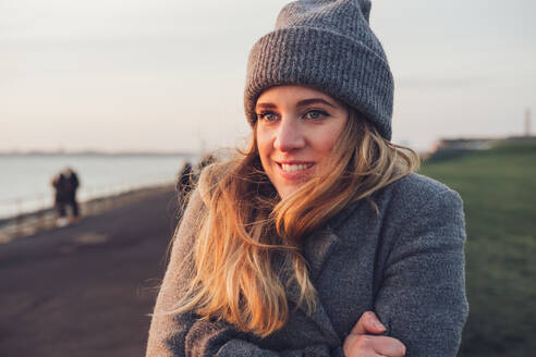 Fröhliche junge Frau in stilvoller Strickkleidung und Hut sitzt auf einer verwitterten Holzbank und genießt den sonnigen Herbsttag auf einer Reise in die Natur - ADSF21331