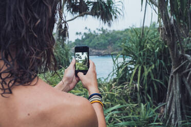 Rückenansicht eines nicht erkennbaren männlichen Reisenden ohne Hemd mit Smartphone, der im grünen Dschungel steht und ein Foto von der Meeresbucht in Hiriketiya in Sri Lanka macht - ADSF21333