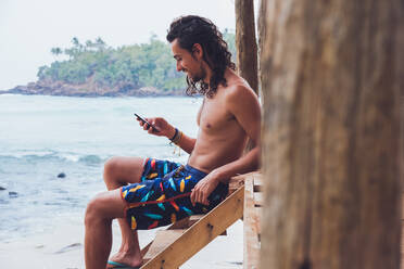 Seitenansicht eines glücklichen männlichen Touristen ohne Hemd, der auf einer Holzterrasse am Meer sitzt und auf seinem Smartphone surft, während er seinen Urlaub in Hiriketiya in Sri Lanka verbringt - ADSF21335