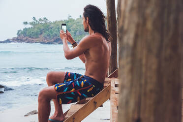 Seitenansicht eines männlichen Touristen ohne Hemd, der auf einer Holzterrasse in der Nähe des Meeres sitzt und mit seinem Smartphone Fotos macht, während er in Hiriketiya in Sri Lanka Urlaub macht - ADSF21336