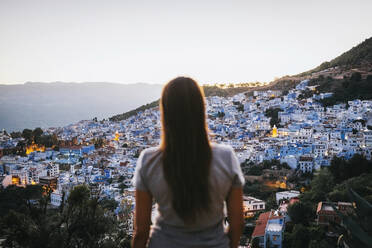 Rückenansicht von unerkennbaren jungen weiblichen Touristen bewundern malerische Stadtbild mit gemütlichen typischen weißen und blauen Häusern auf Hügel Hang gegen Sonnenuntergang Himmel in Chefchaouen gelegen - ADSF21342