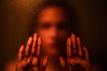 Anonyme, verschwommene Frau, die ein Glas berührt, während sie in einem dunklen Raum mit oranger Beleuchtung steht - ADSF21344