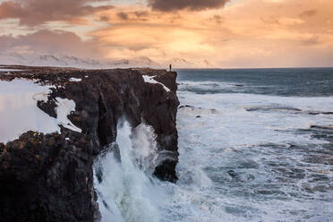 Entfernte Ansicht des Forschers, der auf einer felsigen Klippe steht und die Landschaft des stürmischen Meeres unter dem Sonnenuntergangshimmel in Island bewundert - ADSF21358