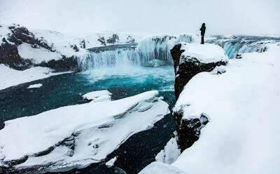 Entfernte Ansicht eines Forschers, der am Rand einer Klippe steht und die Landschaft eines Wasserfalls im Winter in Island bewundert - ADSF21359