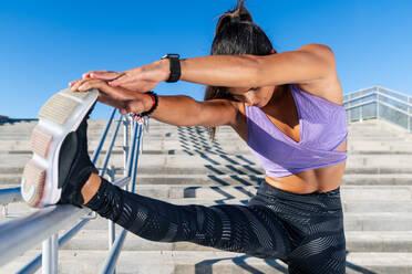 Flexible schlanke Frau in Sportkleidung lehnt sich an das Treppengeländer und streckt die Beine, während sie sich aufwärmt und eine Vorwärtsbeugeübung während des Workouts in der Stadt macht - ADSF21364