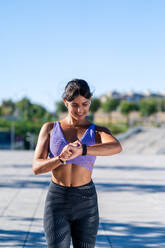 Lächelnde Sportlerin in Sportkleidung, die beim Training in der Stadt an einem sonnigen Tag den Puls auf ihrer Smartwatch überprüft - ADSF21367