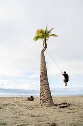 Back view of unrecognizable male swinging on rope hanging from palm tree on beach in Australia - ADSF21425