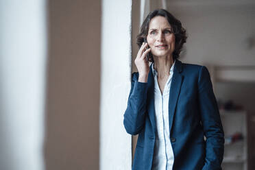 Businesswoman talking on smart phone while leaning on wall at home office - MOEF03665