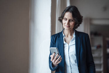 Businesswoman holding smart phone while leaning on wall at home office - MOEF03669