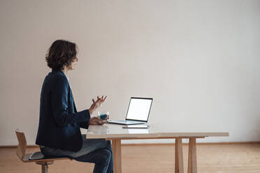 Frau gestikuliert bei der Verwendung eines Laptops auf dem Tisch im Büro zu Hause - MOEF03678