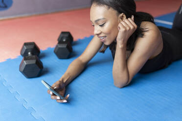 Lächelnde junge Frau, die ein Mobiltelefon benutzt, während sie im Fitnessstudio liegt - JPTF00712