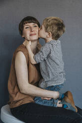 Cute boy kissing mother on cheek over gray background - MFF07625