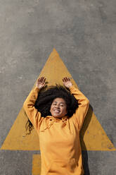 Smiling young woman with arms raised lying on footpath - TCEF01657