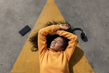 Carefree young woman lying down amidst mobile phone and headphones on footpath - TCEF01660