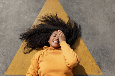 Curly haired woman covering one eye while lying down on footpath - TCEF01662