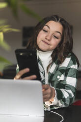 Smiling girl using mobile phone at table in home - JAQF00369