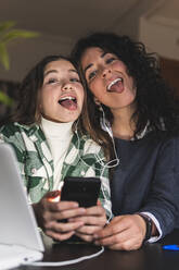Mother and daughter with mobile phone sticking out tongue while listening music through headphones at home - JAQF00372