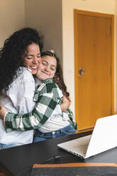 Smiling mother embracing daughter in front of laptop at home - JAQF00379