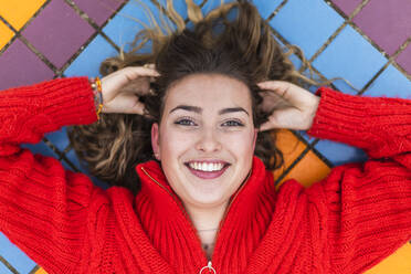 Smiling woman with hand in hair lying on tiled floor - JRVF00353