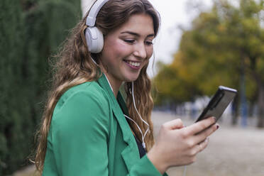 Junge Frau benutzt ihr Smartphone, während sie im Park über Kopfhörer Musik hört - JRVF00363