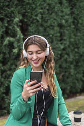 Lächelnde Frau, die ein Smartphone benutzt, während sie im Park über Kopfhörer Musik hört - JRVF00365