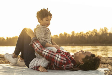 Playful mother and son looking at each other by lake during sunset - UUF22981