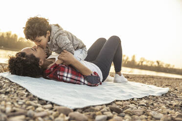 Boy kissing mother lying on blanket over pebbles during sunset - UUF22984