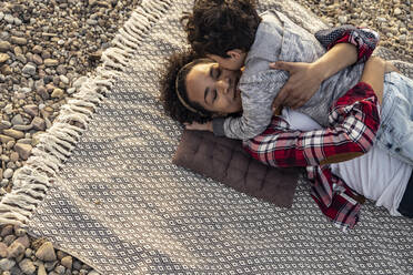 Frau, die einen Jungen umarmt, während sie auf einer Decke über einem Kieselstein liegt - UUF22985