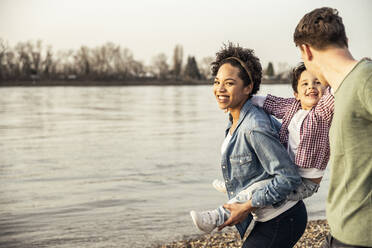 Smiling mother piggybacking son while walking with father by lake - UUF22998