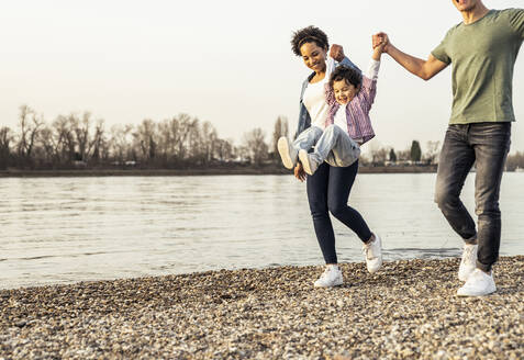 Cheerful boy swinging with parents support over pebble by lake - UUF23003